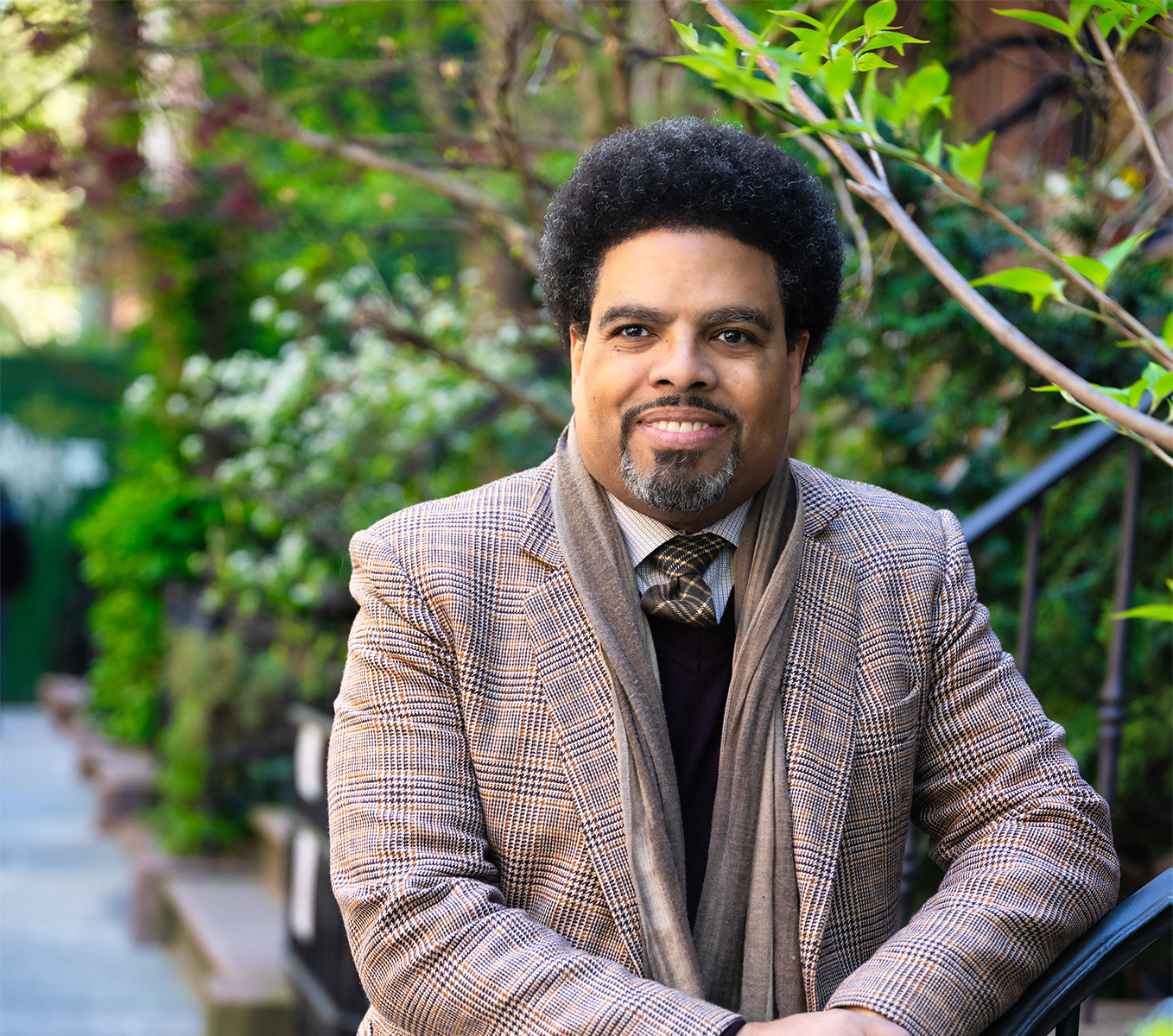 Darrick Hamilton stands in a plaid blazer and scarf leaning on an outdoor stair railing, surrounded by greenery.